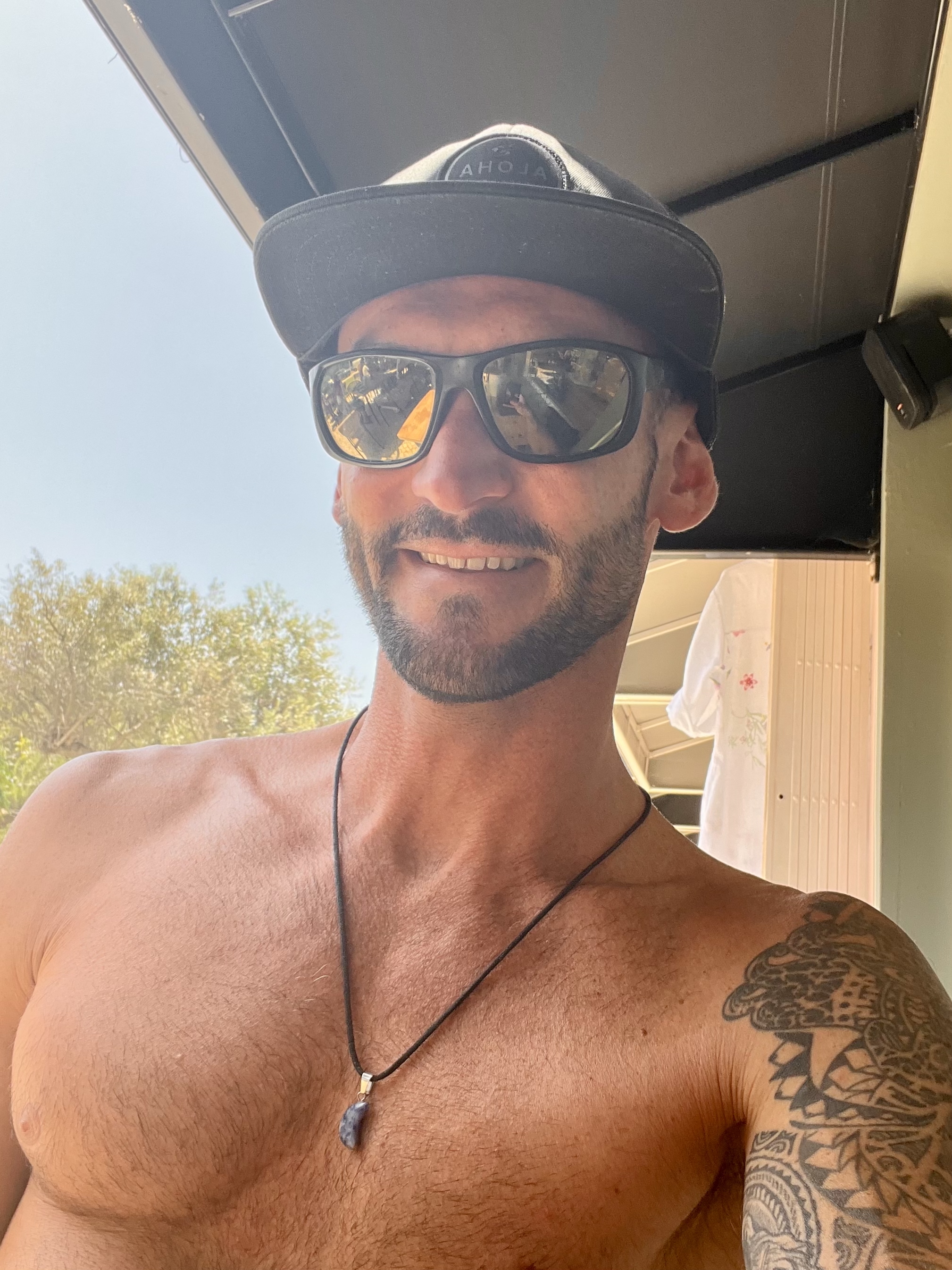 Man wearing a black cap with the word ALOHA, sunglasses, and a necklace with a stone pendant, sitting shirtless outdoors under a shaded structure; tattoo visible on his right shoulder and arm; background includes trees, blue sky, and a white shirt hanging inside a building; relaxed and casual atmosphere